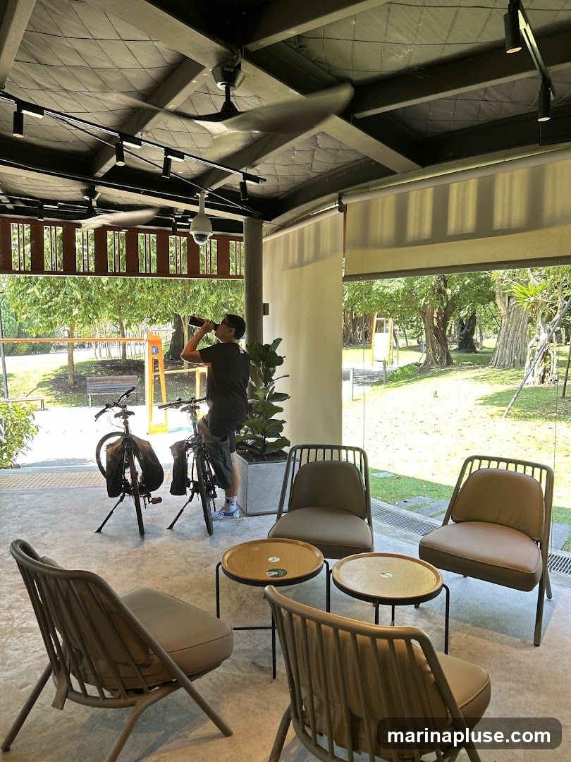 An open-air seating area with modern furniture, including chairs and small tables. A person is standing and drinking from a bottle, with two bicycles parked nearby. The area overlooks a lush green outdoor space with trees and sunlight filtering through. Ceiling fans and track lighting are visible overhead.