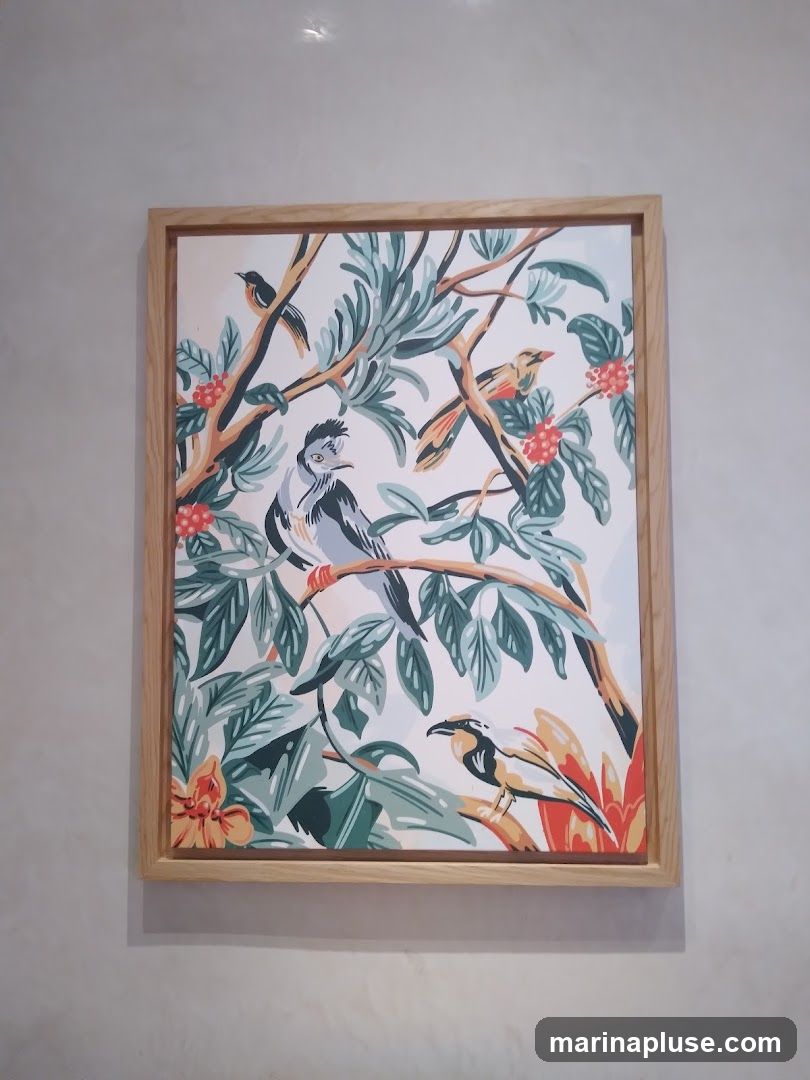 A framed artwork featuring a stylized depiction of birds perched on branches amidst lush foliage and red berries. The artwork has a light-colored wooden frame and is displayed against a textured, light-colored wall.