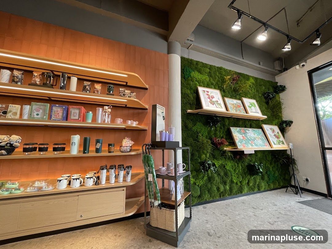 The image shows the interior of a store with shelves displaying various merchandise, including coffee beans, mugs, tumblers, and other accessories. A prominent feature is a large green wall covered in moss and plants, with several framed artworks displayed on shelves in front of it. The overall aesthetic is modern and natural.