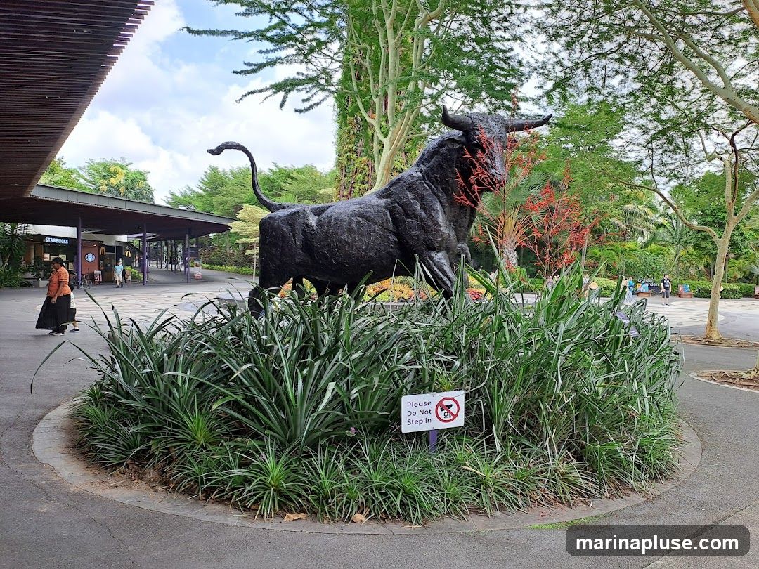 A large, dark bull statue stands prominently in a garden setting, surrounded by lush greenery and flowering plants. In the background, a covered walkway with visible Starbucks and McDonald's signage suggests a commercial area within the garden. A sign near the statue reads "Please Do Not Step In."