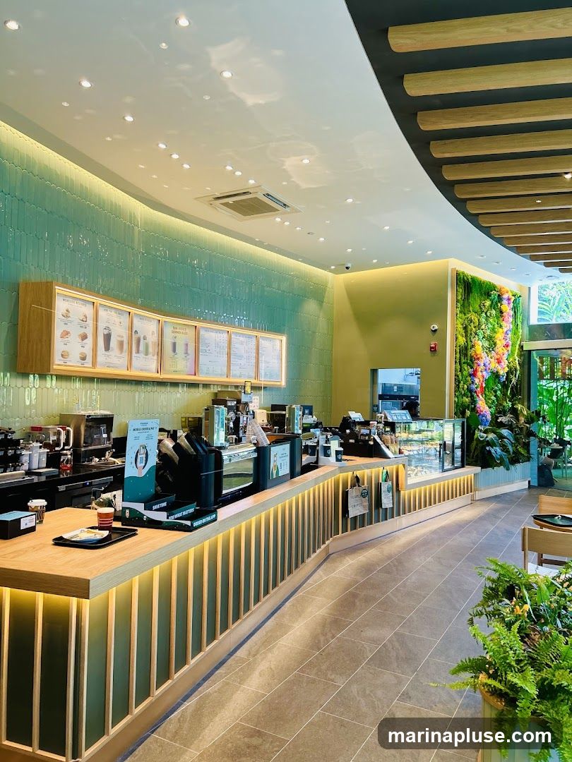 A bright and modern cafe interior featuring a long wooden counter with illuminated vertical slats. Behind the counter, menu boards display various drinks and food items. The walls are decorated with green tiles and a vertical garden with colorful flowers. Seating areas are visible in the background.