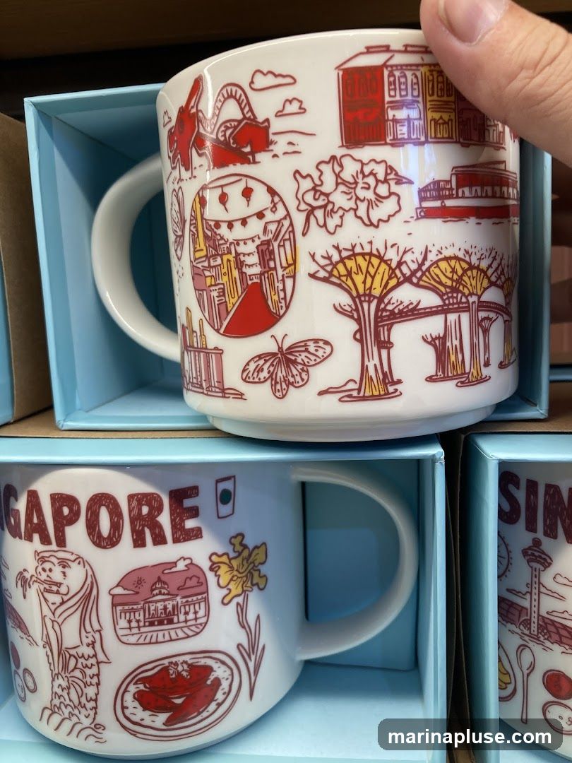 Two white ceramic mugs with red and yellow line drawings of Singapore landmarks and motifs are displayed in light blue gift boxes. The top mug features illustrations of buildings, a dragon, flowers, butterflies, and the Supertrees from Gardens by the Bay. The bottom mug has the word "SINGAPORE" printed in large red letters, along with drawings of the Merlion, a building, a flower, and a dish of food.