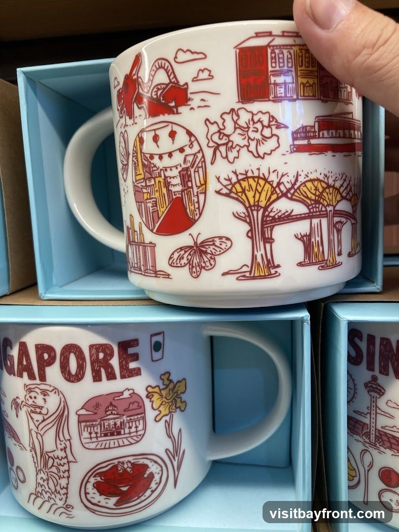 Two white ceramic mugs with red and yellow line drawings of Singapore landmarks and motifs are displayed in light blue gift boxes. The top mug features illustrations of buildings, a dragon, flowers, butterflies, and the Supertrees from Gardens by the Bay. The bottom mug has the word "SINGAPORE" printed in large red letters, along with drawings of the Merlion, a building, a flower, and a dish of food.