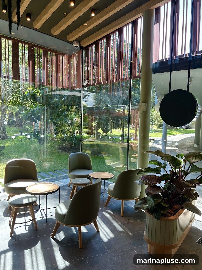 A contemporary cafe interior features a seating area with several plush, olive-green armchairs and small wooden tables. Large glass windows offer a view of lush greenery and trees outside. The ceiling has a modern design with exposed wooden beams and track lighting. A large potted plant adds a touch of nature to the space. A circular black sign hangs from the ceiling, its purpose unclear.