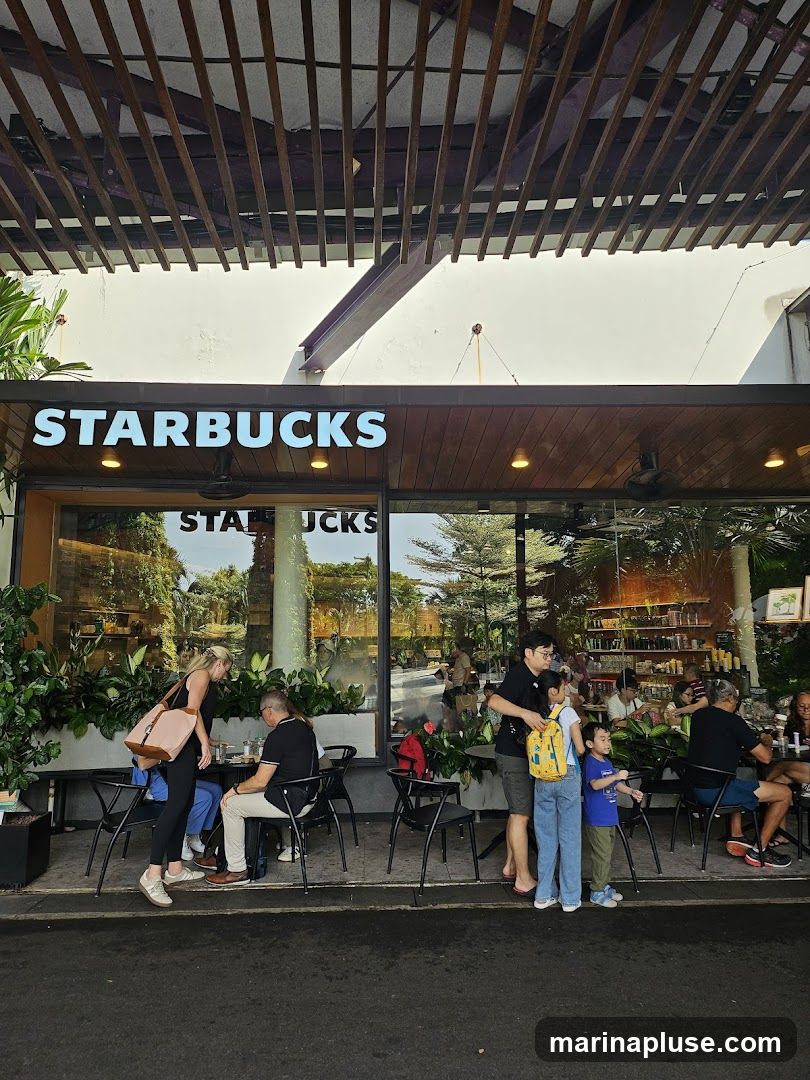 The exterior of a Starbucks coffee shop with large glass windows and a prominent Starbucks sign above the entrance. People are seated at tables outside the shop, and others are standing near the entrance. The shop is decorated with plants.
