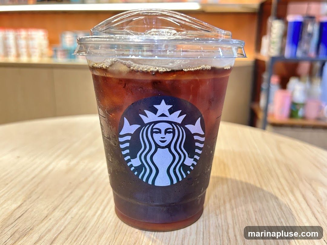A close-up shot of an iced coffee in a clear plastic cup with the Starbucks logo visible. The drink is filled with ice and dark liquid, and the cup has a clear plastic lid. The cup sits on a light-colored wooden table. In the blurred background, shelves with various items and an orange-colored wall are visible.