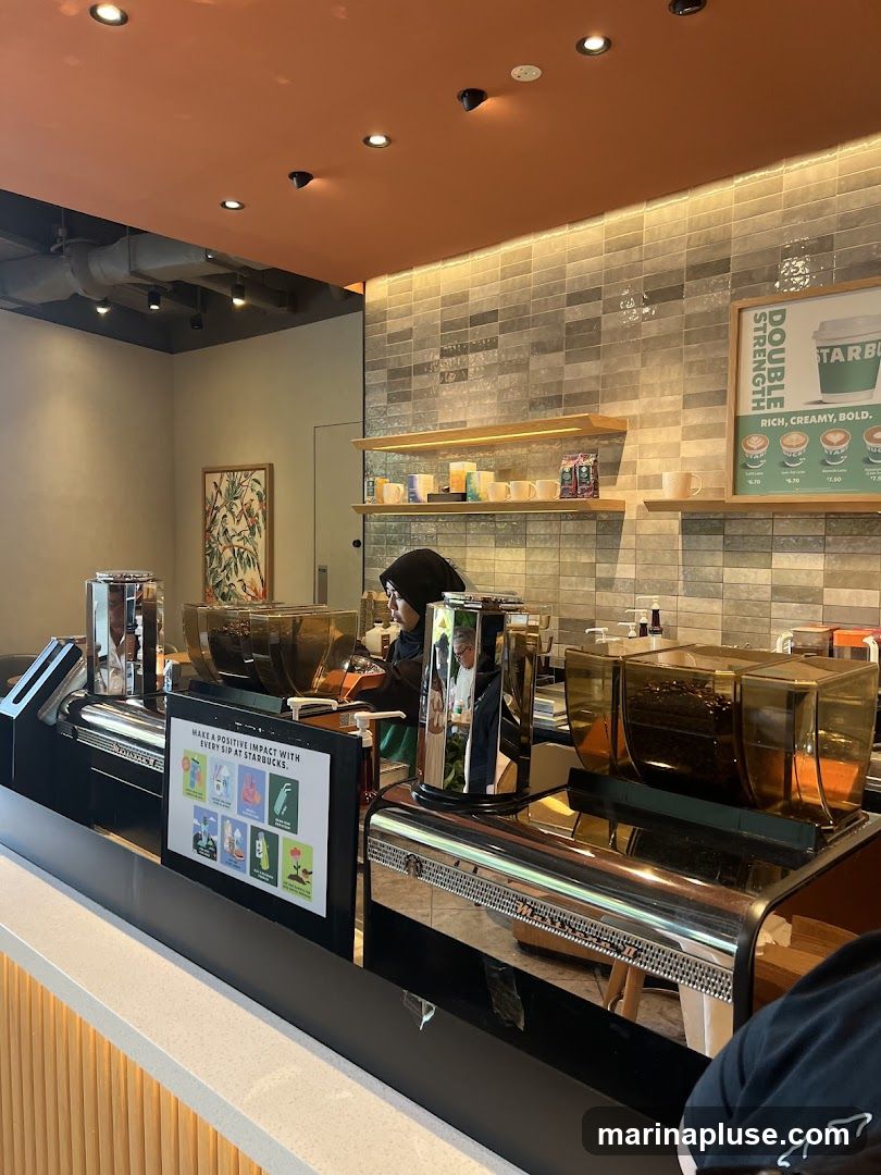 A view of a Starbucks counter with coffee machines and a barista. There is a sign that says "MAKE A POSITIVE IMPACT WITH EVERY SIP AT STARBUCKS." Another sign displays "DOUBLE STRENGTH RICH, CREAMY, BOLD." Shelves behind the counter hold mugs and coffee bags.