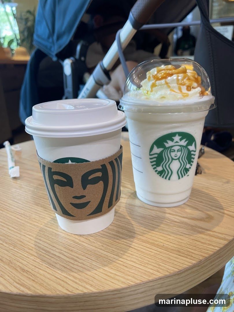 Two Starbucks drinks are on a wooden table. One is a white cup with a lid and a brown sleeve featuring the Starbucks logo. The other is a clear plastic cup with a dome lid, filled with a white beverage topped with whipped cream and caramel drizzle, also featuring the Starbucks logo. A stroller is visible in the background.