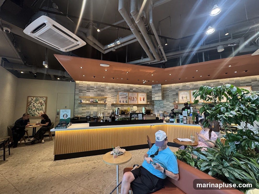 An interior view of a coffee shop with customers seated at tables and at the counter. The counter area features a display of drinks and pastries, with baristas preparing orders. There are also plants and artwork decorating the space.