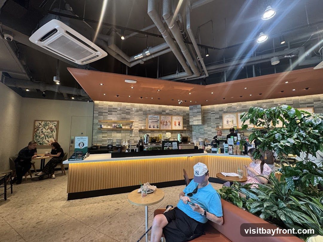 An interior view of a coffee shop with customers seated at tables and at the counter. The counter area features a display of drinks and pastries, with baristas preparing orders. There are also plants and artwork decorating the space.