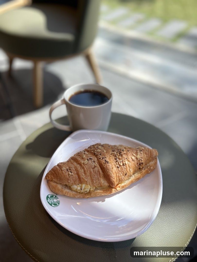 A croissant filled with egg salad and topped with seeds sits on a white plate with a Starbucks logo. A white mug of black coffee is next to the plate. The setting appears to be indoors with natural light streaming in, casting shadows on the table. A blurred green chair is visible in the background.