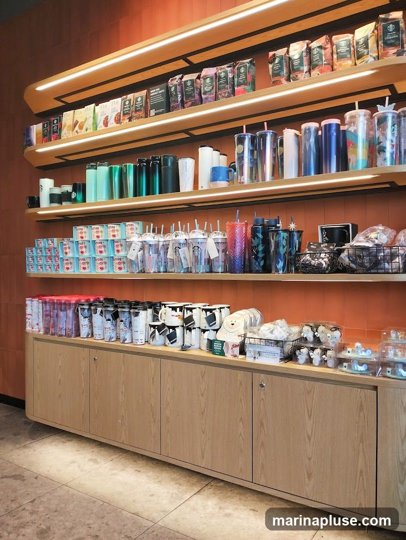 Shelves stocked with Starbucks coffee bags, tumblers, mugs, and other merchandise. The display features a variety of colorful tumblers, reusable cups, and coffee bean packages. Below, a wooden cabinet displays more merchandise, including mugs, small plush toys, and other gift items.