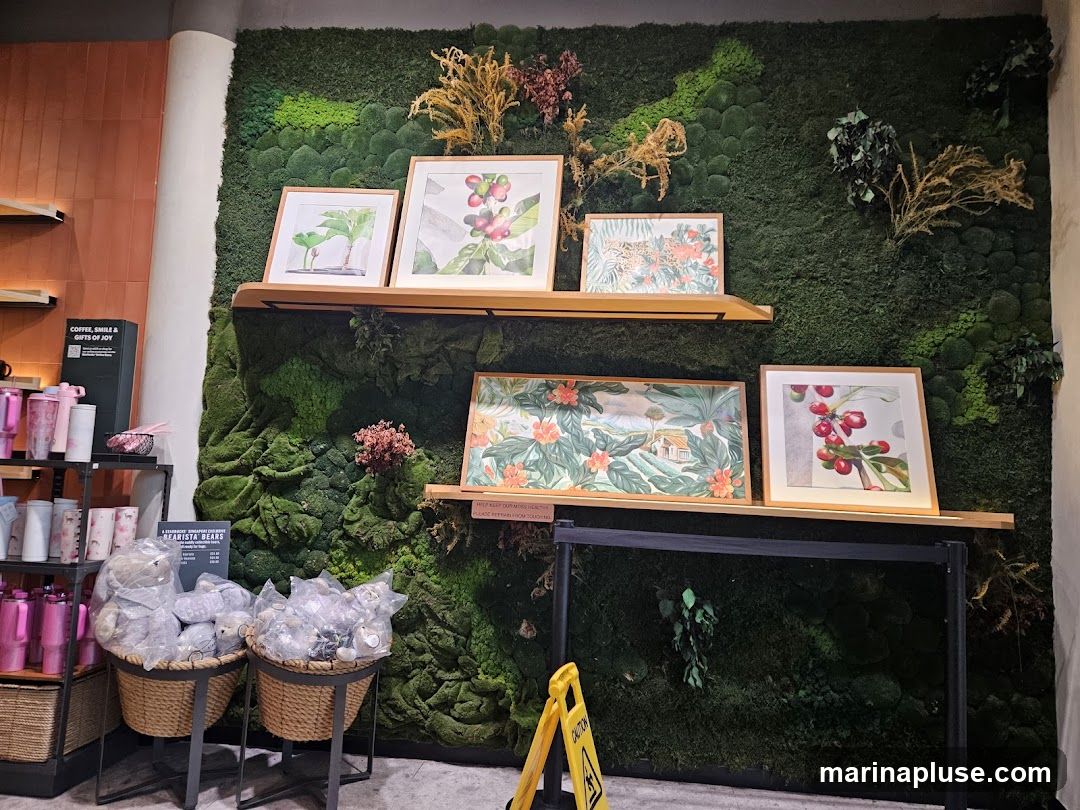 A wall covered in a lush green moss installation serves as a backdrop for several framed artworks. The artwork depicts botanical illustrations of coffee plants, a leopard in a floral setting, and a tropical landscape. Below the shelves displaying the art, two wicker baskets hold plush teddy bears in plastic packaging. To the left, shelves display various pink and white tumblers and mugs. A sign reads "COFFEE, SMILE & GIFTS OF JOY". A yellow caution sign is visible in the lower right corner.