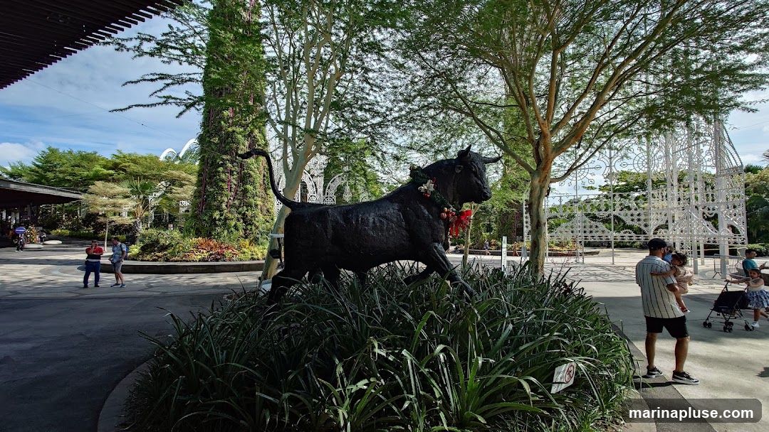 A large, dark statue of a bull stands prominently in a lush garden setting. The bull is adorned with a festive garland around its neck. In the background, there are tall trees, a large green structure covered in plants, and an intricate white lattice structure. Several people are visible in the scene, including a man holding a child and other families enjoying the outdoor space. The sky is partly cloudy.