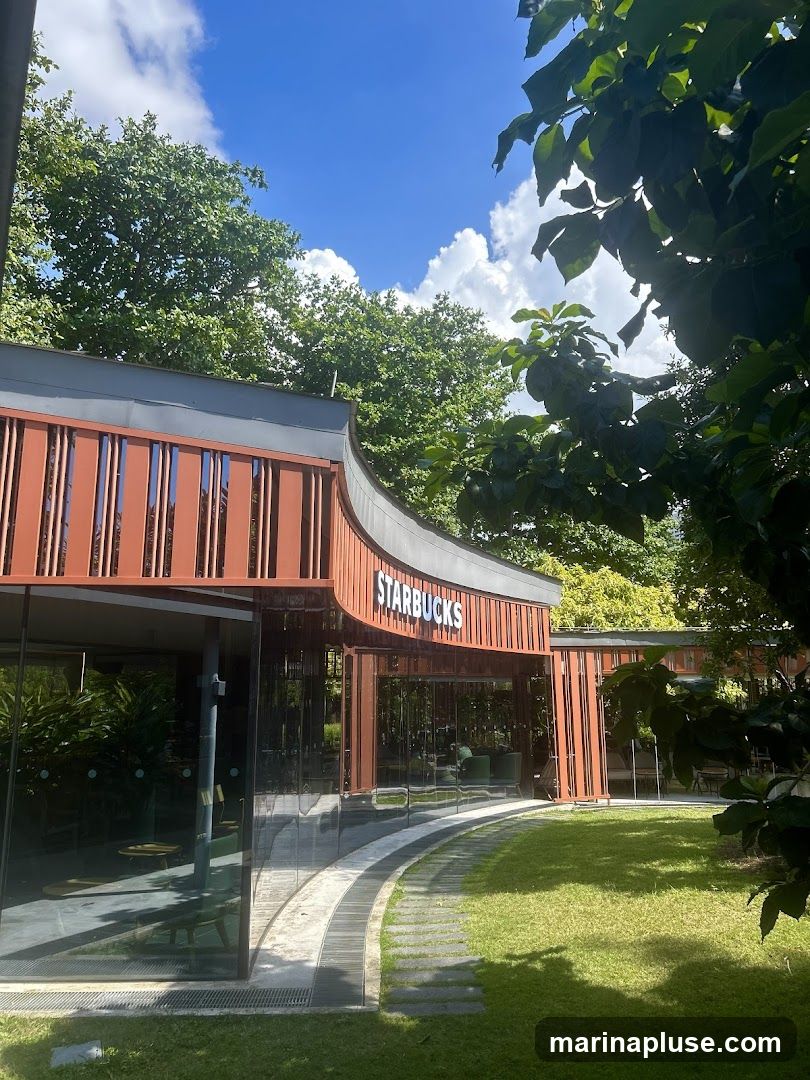 A modern Starbucks store with a curved wooden facade and large glass windows. The Starbucks logo is prominently displayed above the entrance. The store is surrounded by lush green grass and trees, with a pathway leading to the entrance.