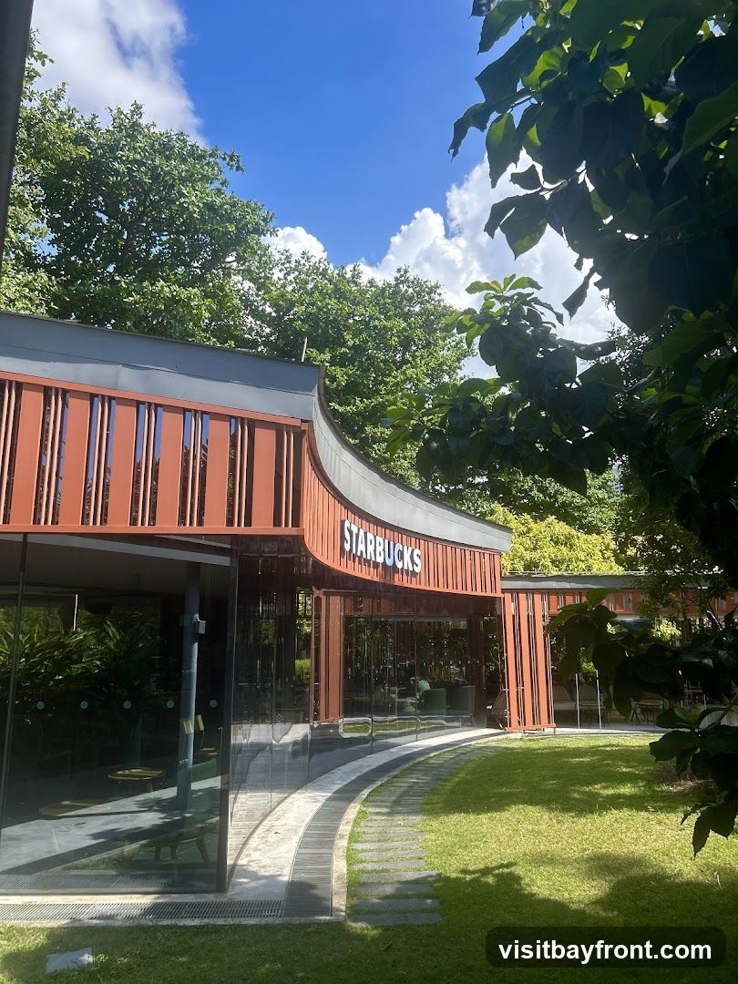 A modern Starbucks store with a curved wooden facade and large glass windows. The Starbucks logo is prominently displayed above the entrance. The store is surrounded by lush green grass and trees, with a pathway leading to the entrance.