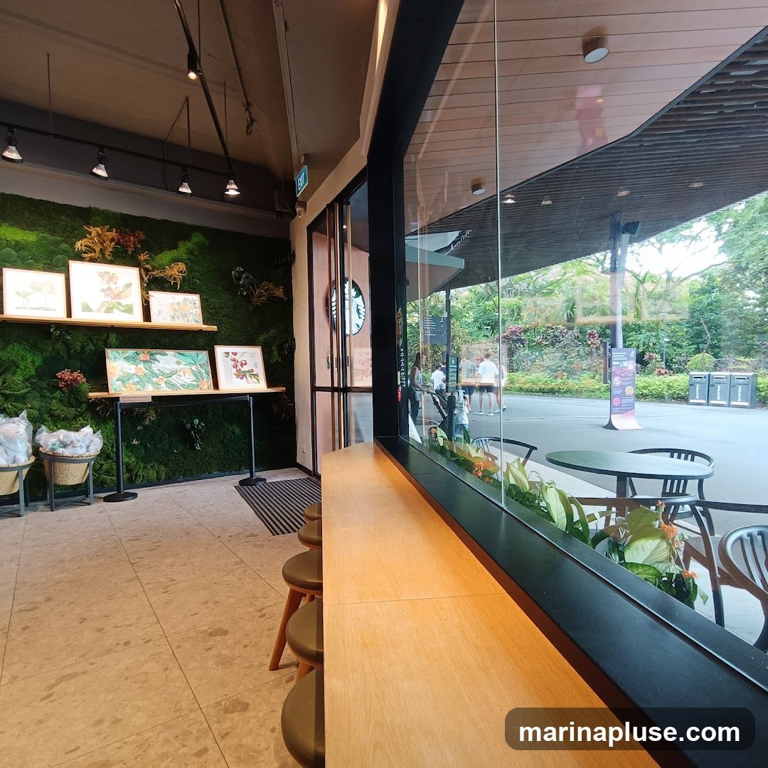 The image shows the interior of a Starbucks cafe, with a prominent green moss wall adorned with framed artwork. A wooden counter with stools runs along the window, offering a view of the outdoor seating area and lush greenery. The Starbucks logo is visible on the glass doors.