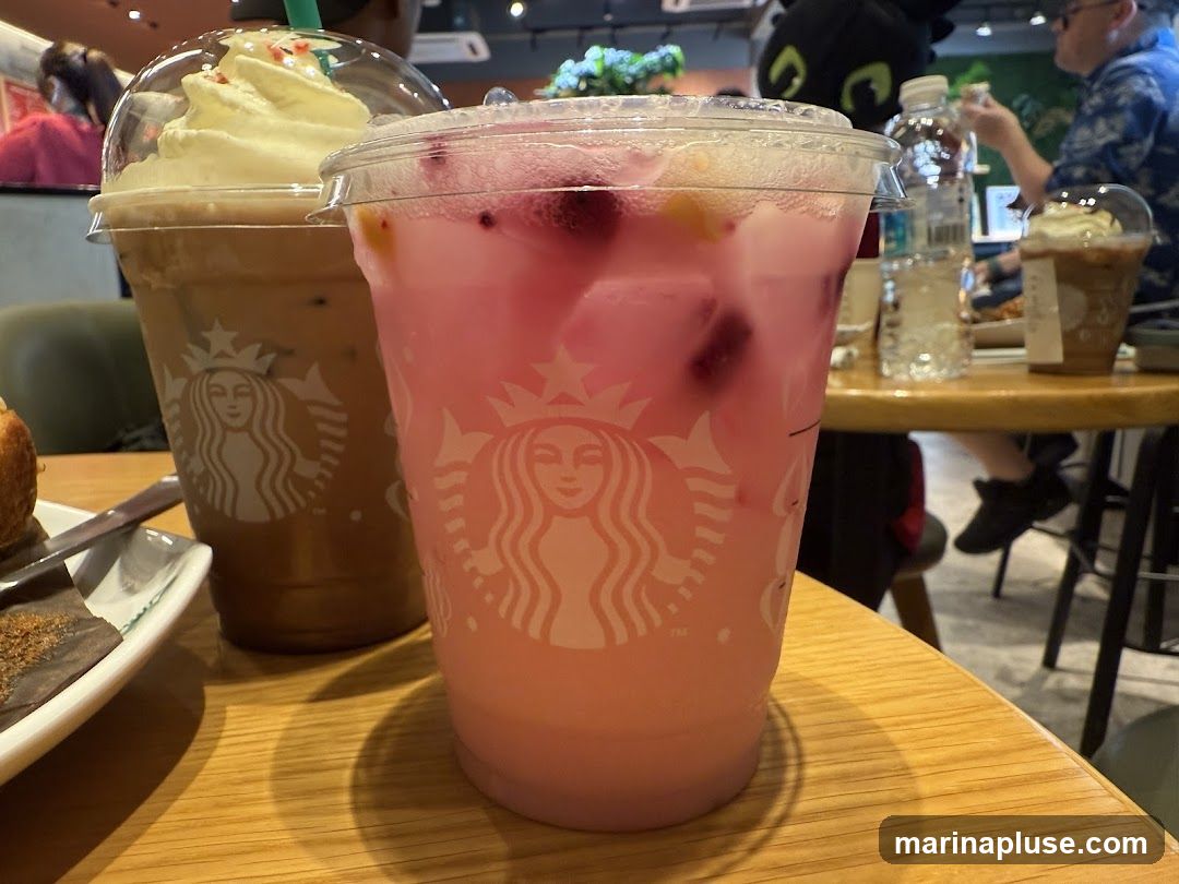 Two Starbucks drinks are on a wooden table. The drink in the foreground is pink with berries and ice. The drink in the background is brown with whipped cream. Other patrons and a bottle of water are visible in the background.