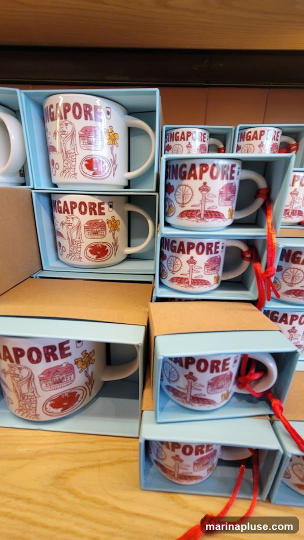 A display of white ceramic mugs with red illustrations and the word "SINGAPORE" printed on them. The mugs are packaged in light blue boxes with cardboard inserts. The illustrations depict landmarks and symbols associated with Singapore, such as the Merlion, a Ferris wheel, and various buildings. Some mugs are larger, while others are smaller, possibly espresso cups. Red strings with tassels are attached to some of the smaller mugs. The display is set against a wooden surface.