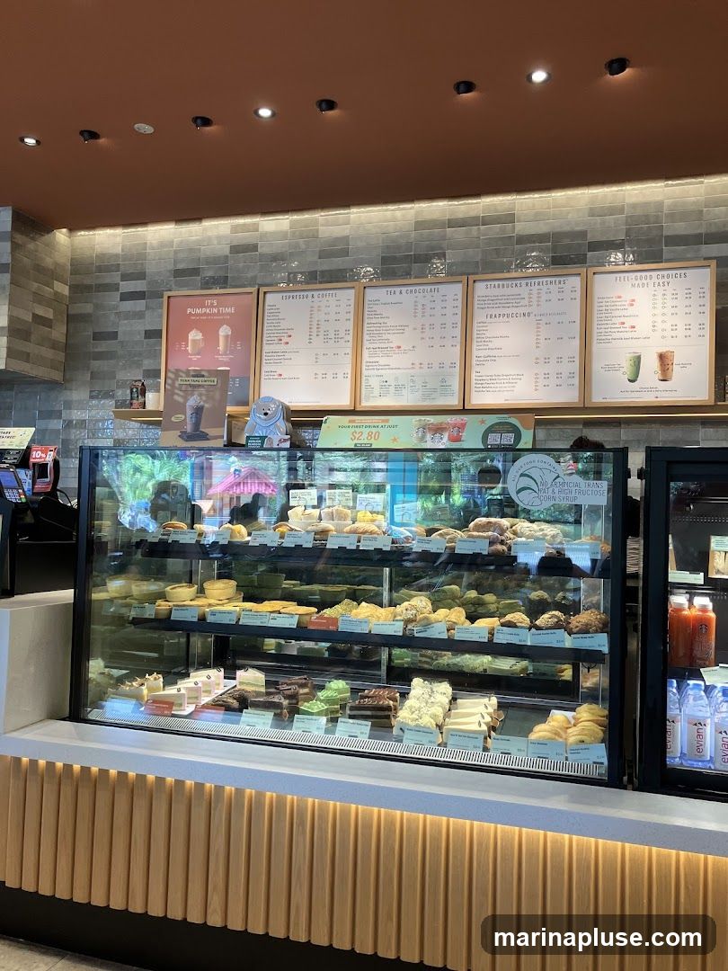 A display case filled with various pastries and desserts, including cakes, tarts, and buns. Above the display are menu boards listing coffee, tea, and other beverages. Bottles of water and juice are visible to the right.