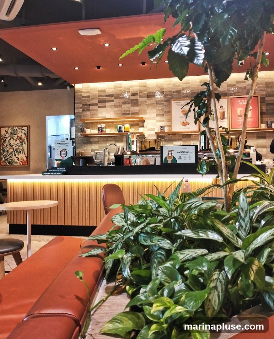 The image shows the interior of a modern cafe. In the foreground, there is a long, burnt orange sofa and lush green plants. A small round table with a stool is visible to the left. The background features a counter with coffee machines and shelves stocked with cups and other items. Two framed posters are displayed on the tiled wall behind the counter. A digital screen on the counter displays a message about ordering on the app. The overall ambiance is clean and contemporary, with warm lighting and natural elements.