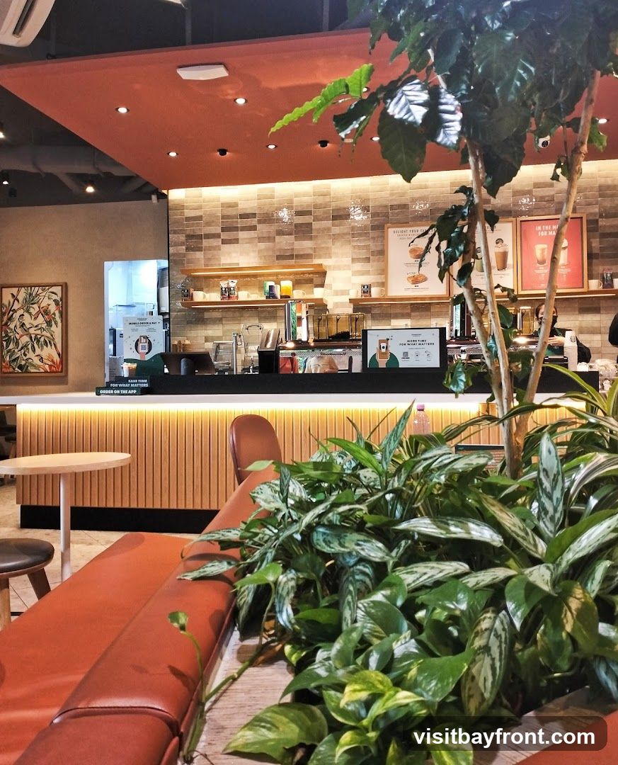 The image shows the interior of a modern cafe. In the foreground, there is a long, burnt orange sofa and lush green plants. A small round table with a stool is visible to the left. The background features a counter with coffee machines and shelves stocked with cups and other items. Two framed posters are displayed on the tiled wall behind the counter. A digital screen on the counter displays a message about ordering on the app. The overall ambiance is clean and contemporary, with warm lighting and natural elements.