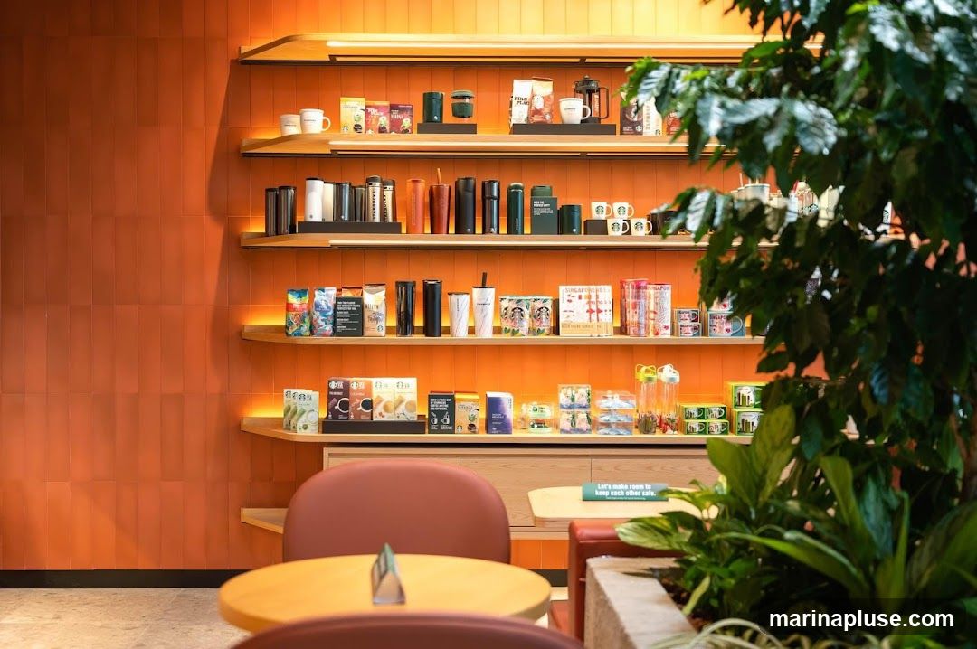 Shelves filled with Starbucks branded merchandise, including tumblers, mugs, coffee beans, and other accessories, against an orange tiled wall. A lush green plant is in the foreground.