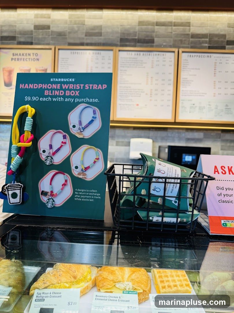 A display at Starbucks featuring a promotion for Handphone Wrist Strap Blind Boxes, priced at $9.90 with any purchase. The display includes a poster showing the product and a basket filled with the blind boxes. In the foreground, a refrigerated display case shows various food items, including croissants and a wafflewich. Menu boards are visible in the background.