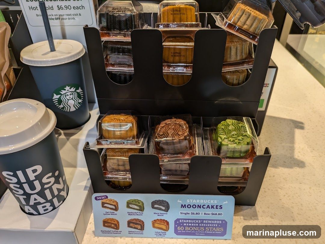 A display of Starbucks mooncakes and reusable cups. The mooncakes are individually packaged and arranged in a black display stand. Several flavors are visible, including coffee, matcha, and what appears to be a Starbucks logo imprinted on one. Two reusable Starbucks cups are also present, one with the "Sip Sustainably" slogan. A sign details the pricing for single mooncakes and boxes, as well as a Starbucks Rewards promotion.