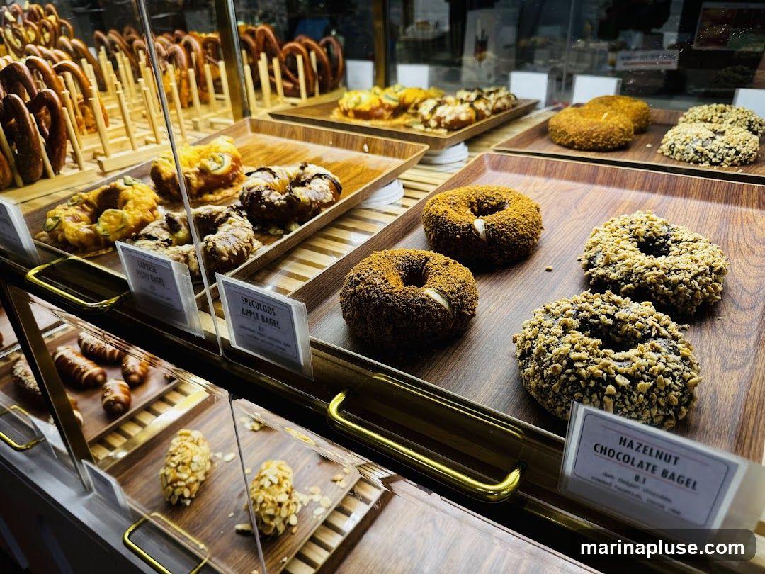 A display case filled with various baked goods, including bagels, pretzels, and other pastries. In the foreground, there are bagels with different toppings, such as nuts and cinnamon. Behind them, there are savory bagels with cheese and what appear to be jalapeños. Further back, rows of pretzels are visible.