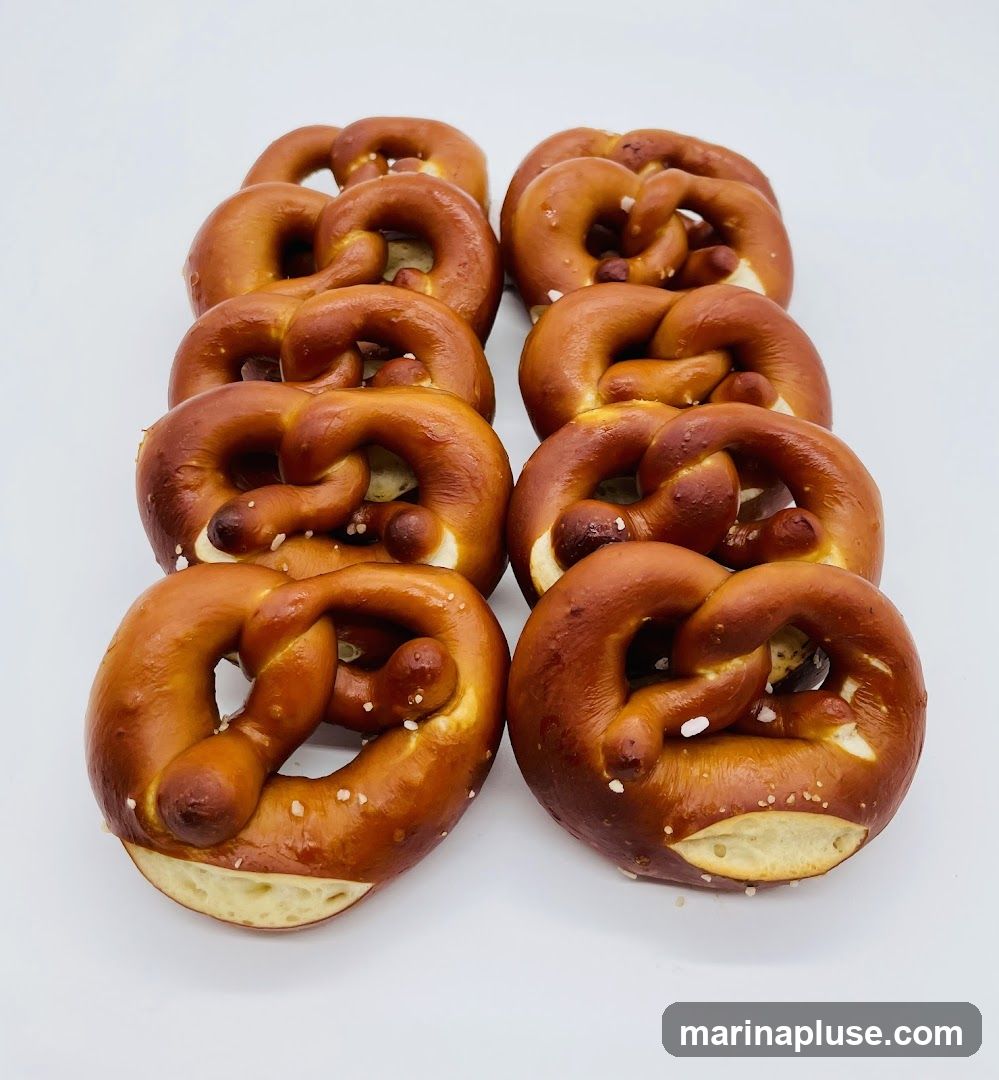 A close-up studio shot of eight freshly baked pretzels arranged in two rows of four against a white background. The pretzels are golden brown with a glossy sheen and sprinkled with coarse salt crystals. Their classic twisted shape is clearly visible, with the dough appearing soft and slightly chewy.