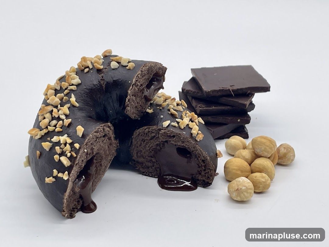 A chocolate donut is cut in half, revealing a rich chocolate filling. The donut is topped with chopped hazelnuts and served with a stack of dark chocolate squares and whole hazelnuts on a white background.