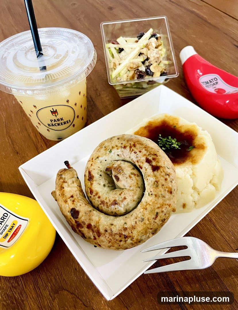 A coiled sausage is served on a white plate with a side of mashed potatoes topped with gravy and a small container of apple and walnut salad. A plastic cup with a beverage and the logo "PARK BÄCKEREI" is visible, along with a bottle of ketchup and a yellow mustard dispenser. A fork rests on the plate.