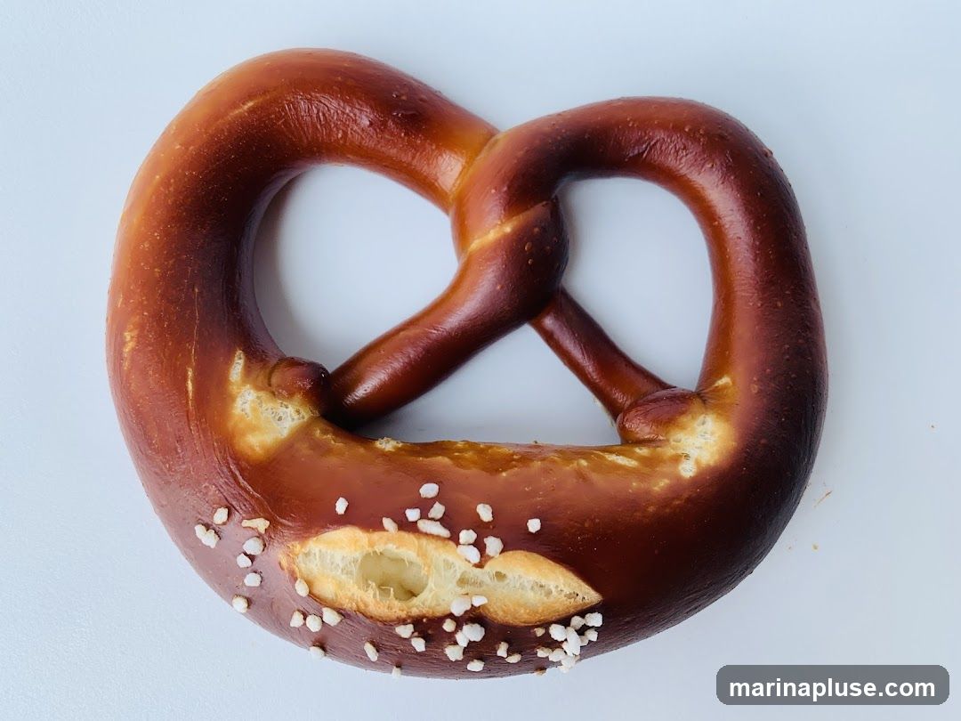 A close-up, top-down view of a freshly baked pretzel with coarse salt sprinkled on its surface. The pretzel has a glossy, golden-brown crust and a soft, doughy interior visible in some areas.