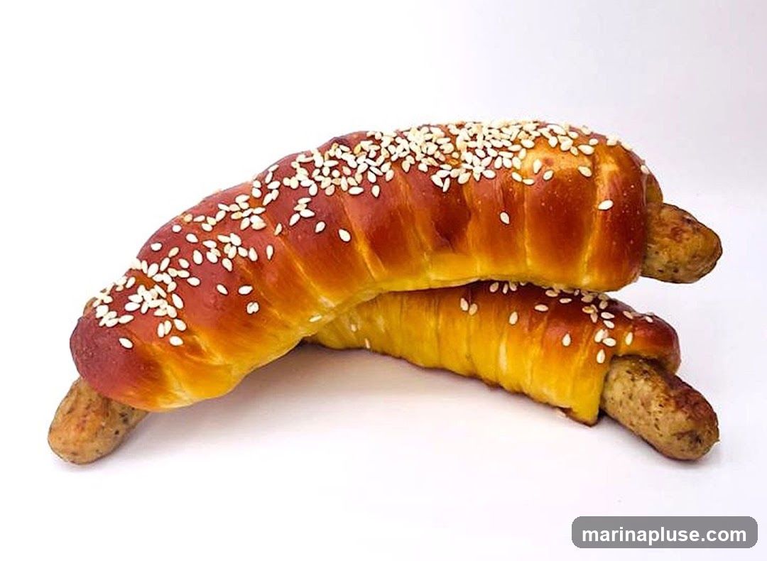 Two sausage rolls, each with a sausage wrapped in a golden-brown, sesame-seed-topped pastry, are stacked on a white background.