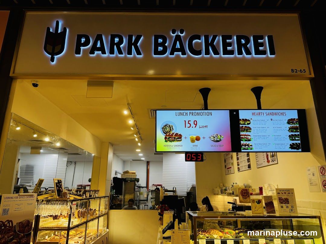 A view of the interior and exterior of a bakery called Park Bäckerei. A large illuminated sign with the bakery's name is visible above the entrance. Digital screens display a lunch promotion and a menu of hearty sandwiches. Display cases filled with baked goods and sandwiches are visible inside the store.