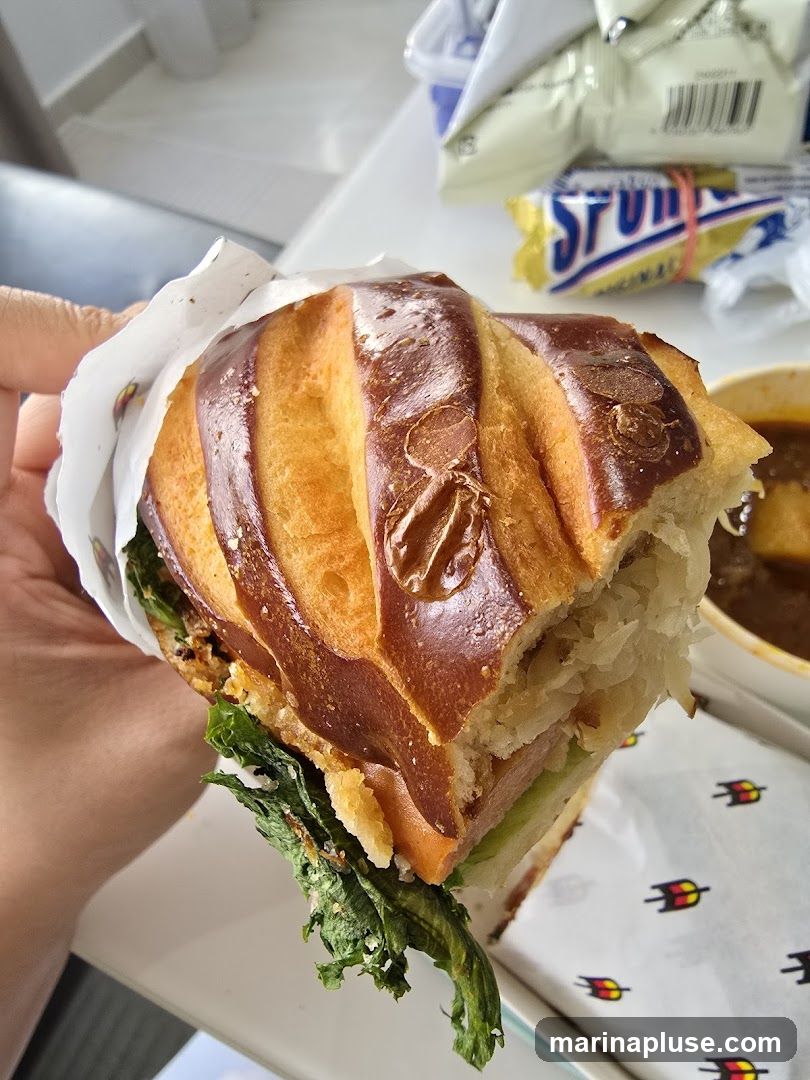 A close-up shot of a person holding a large sandwich with a pretzel-like bun. The sandwich is filled with lettuce, meat, and possibly cheese. In the background, a bowl of soup and some packaged snacks are visible. The wrapper of the sandwich has a repeating logo with three arrows.