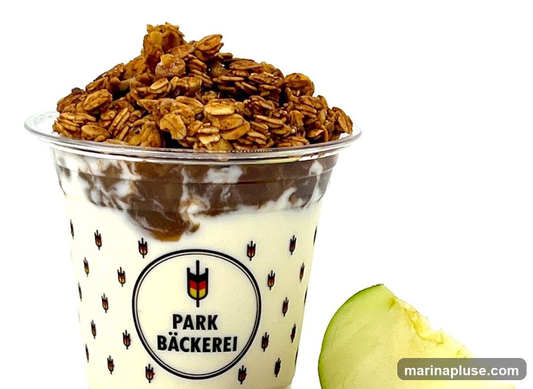 A clear plastic cup filled with layers of yogurt and a dark sauce, topped with a generous portion of granola. The cup is decorated with a repeating pattern of small red tulip-like figures and a central circular logo that reads "PARK BÄCKEREI" with a German flag emblem. A slice of green apple is visible to the right of the cup.