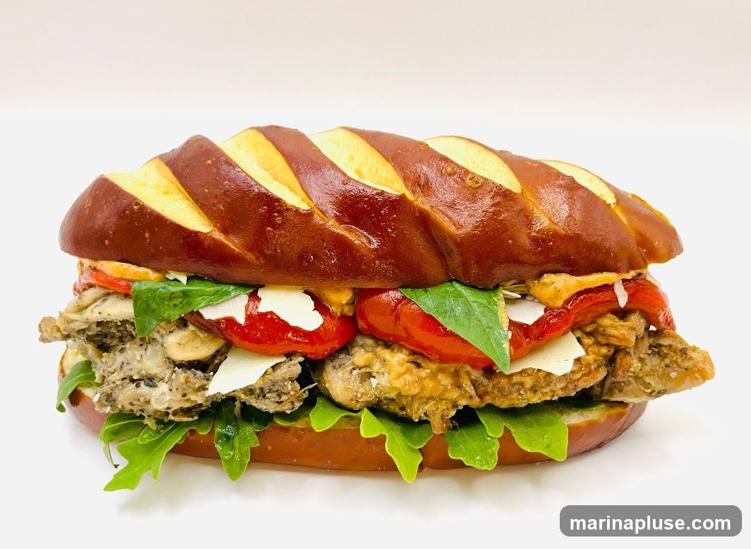 A close-up shot of a chicken sandwich on a pretzel bun. The sandwich is filled with chicken, roasted red peppers, cheese, basil, and arugula.