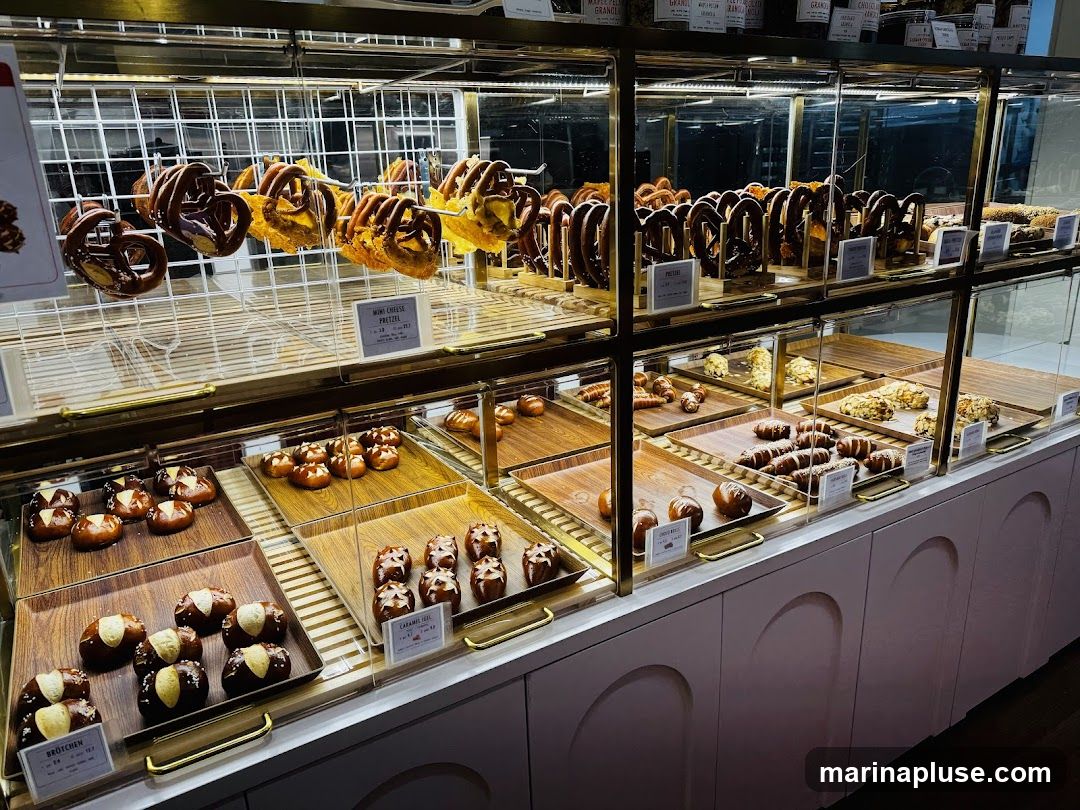 A well-lit bakery display case filled with various pastries, including pretzels, rolls, and other baked goods. The items are arranged on multiple shelves and trays, with some items labeled with small signs.