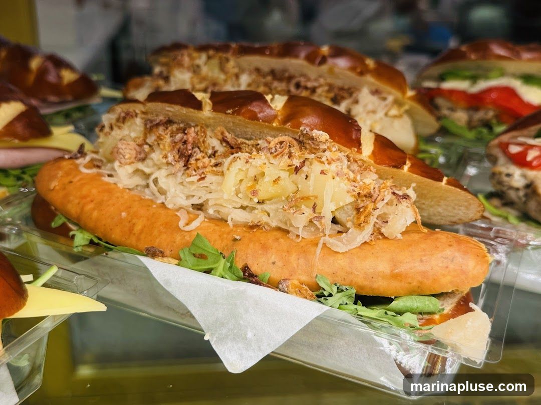 A close-up shot of a sausage sandwich served in a pretzel bun. The sandwich is filled with a large sausage, sauerkraut, and crispy fried onions, with some greens underneath. Other sandwiches are visible in the background.