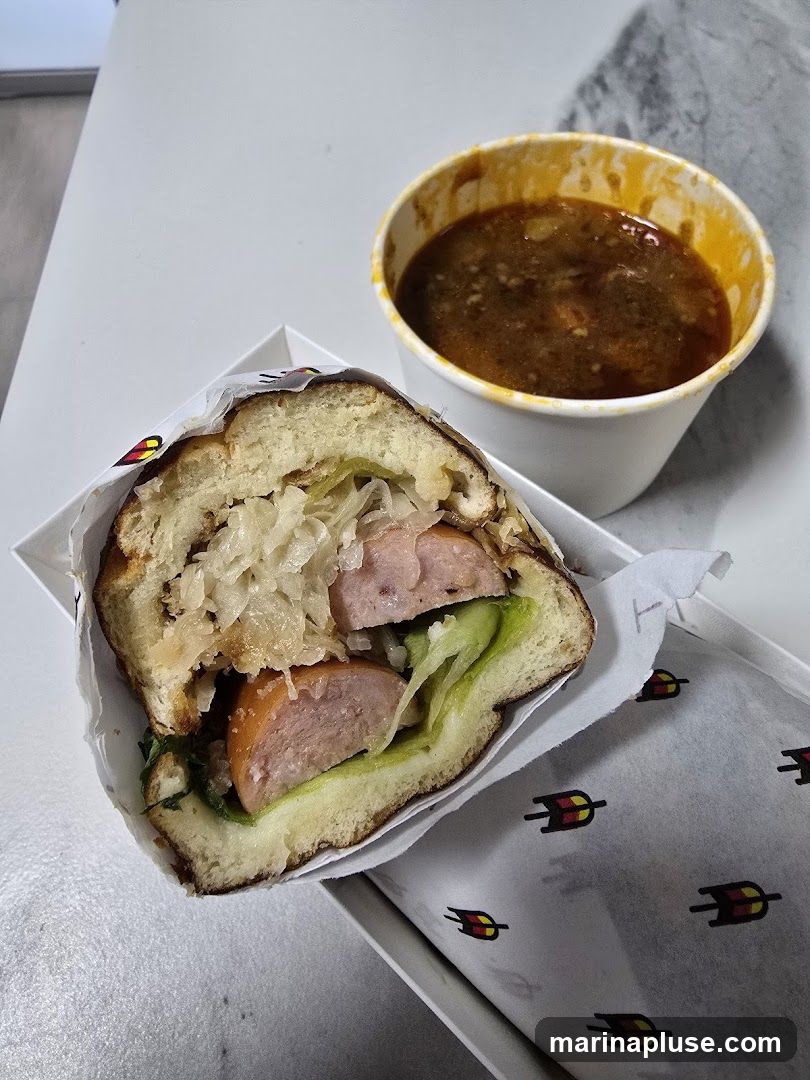 A close-up shot of a sausage sandwich and a cup of soup. The sandwich is on a bun with lettuce, sauerkraut, and a thick sausage. The soup is in a white paper cup with a yellow rim. The food is presented in white paper packaging with a repeating logo.