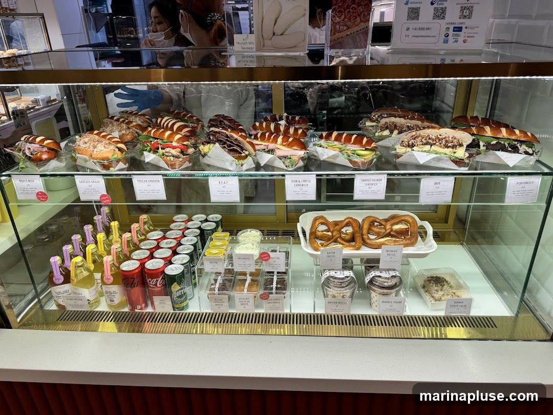 A refrigerated display case filled with various sandwiches, pretzels, drinks, and desserts. The sandwiches are arranged on the top shelf, with labels indicating their fillings. Below the sandwiches are bottles of drinks and cans of soda. The bottom shelf features pretzels, yogurt, and potato salad.