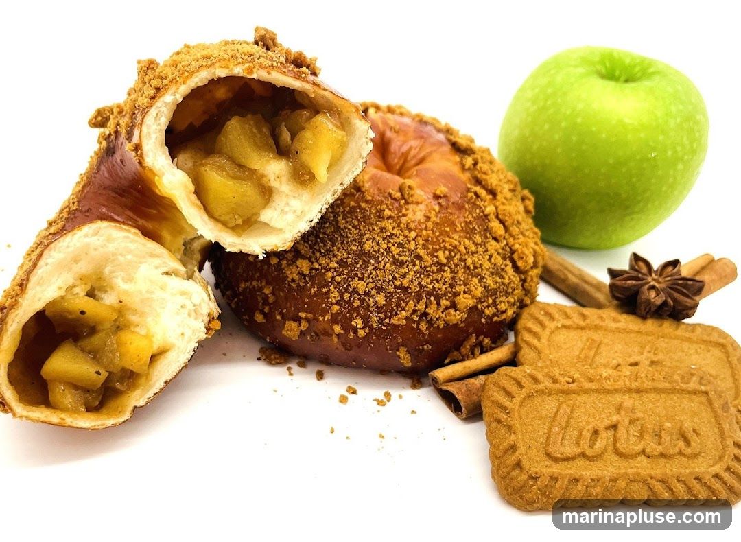 A close-up shot of a pastry filled with apple chunks, coated in a crumb topping. Next to it is a whole pastry, a green apple, cinnamon sticks, star anise, and two Lotus biscuits.