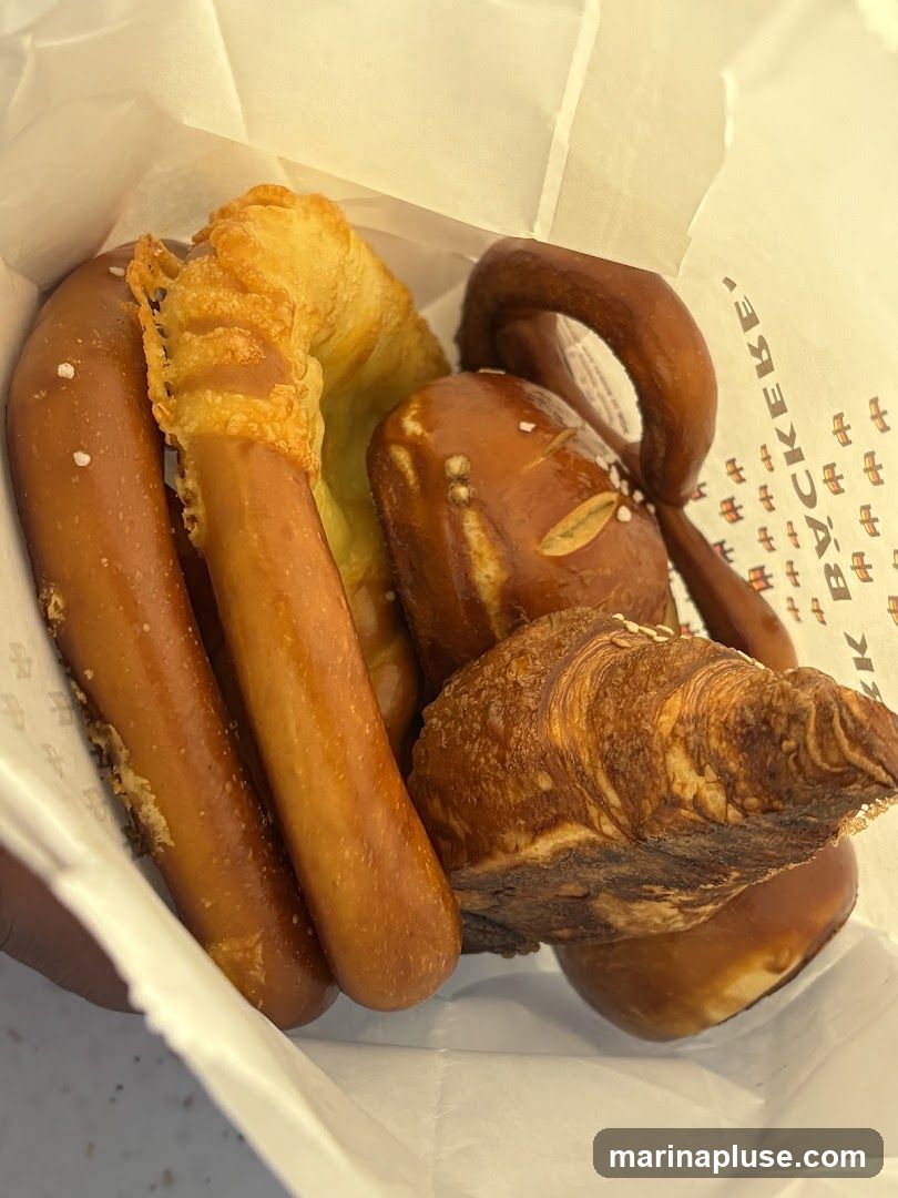 A white paper bag filled with various baked goods, including pretzels, a croissant, and a cheese-covered pastry. The bag has a repeating pattern of Chinese characters and the partial text "К ВУСКEBE'" visible on the inside.