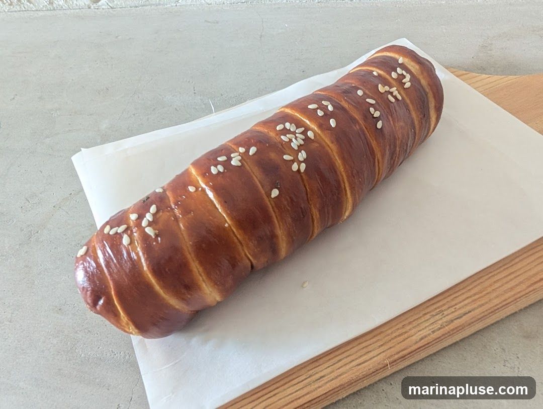A close-up shot of a long, golden-brown pretzel roll topped with sesame seeds. The roll is placed on a piece of white parchment paper, which rests on a wooden board. The background is a textured, light-colored surface.