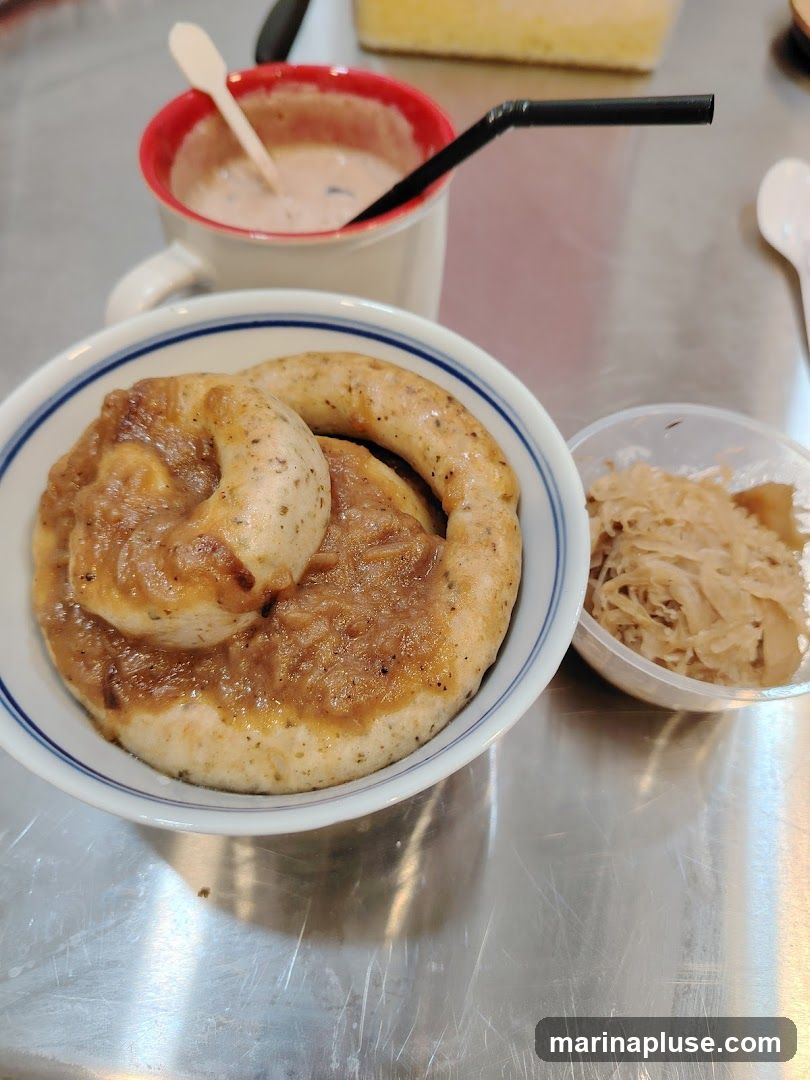 A close-up shot of a meal featuring a large pretzel-shaped sausage covered in gravy, served in a bowl with a side of sauerkraut in a clear plastic container. A mug of a creamy beverage with a straw and a small spoon is also visible. The meal is presented on a metallic surface.