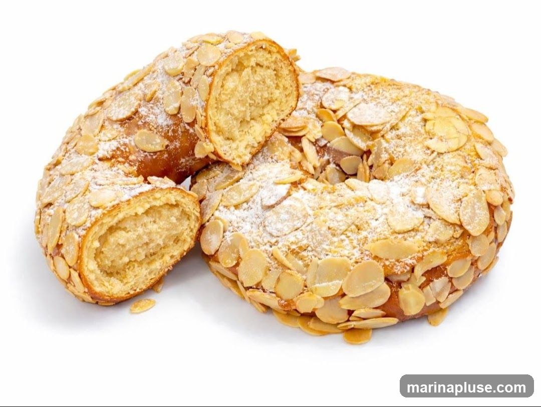 A ring-shaped cake, possibly a Roscón de Reyes, is covered in sliced almonds and dusted with powdered sugar. One section of the cake is cut and separated, revealing a dense, pale filling. The cake is presented on a white background.