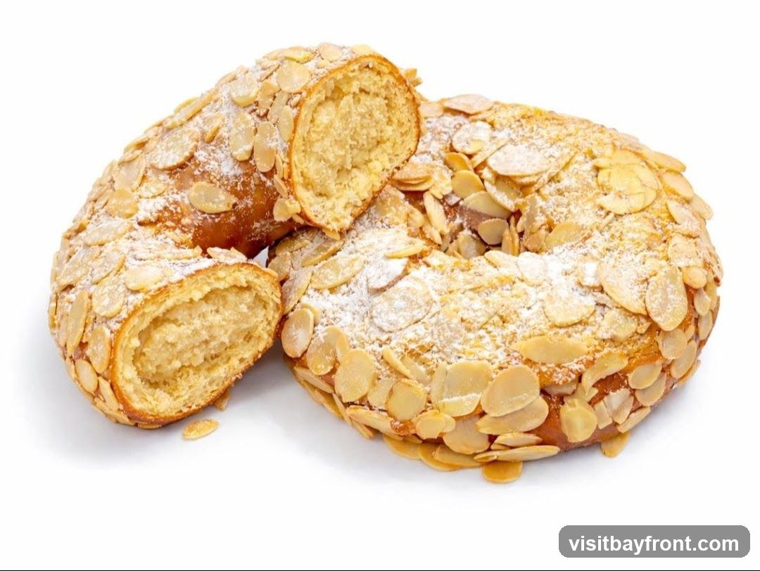 A ring-shaped cake, possibly a Roscón de Reyes, is covered in sliced almonds and dusted with powdered sugar. One section of the cake is cut and separated, revealing a dense, pale filling. The cake is presented on a white background.