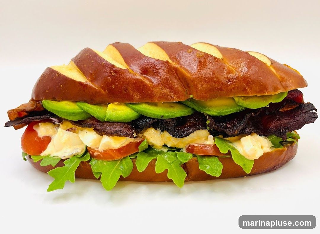 A close-up studio shot of a sandwich on a white background. The sandwich is made with a pretzel bun and filled with sliced avocado, bacon, tomatoes, arugula, and a creamy spread.